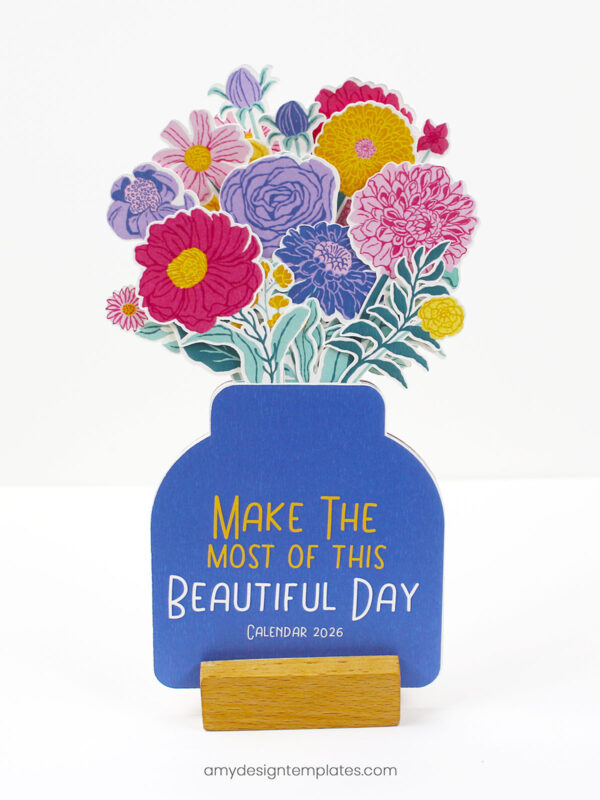 DIY floral desk calendar for 2026 showing a blue jar-shaped card with a "Make the Most of This Beautiful Day" cover, topped with colorful paper-cut flowers and held in a wooden block stand.